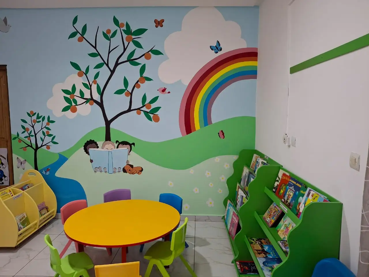 Une table ronde jaune et des chaises d’enfant devant une fresque peinte d’un arbre et d’un arc-en-ciel, dans le coin lecture